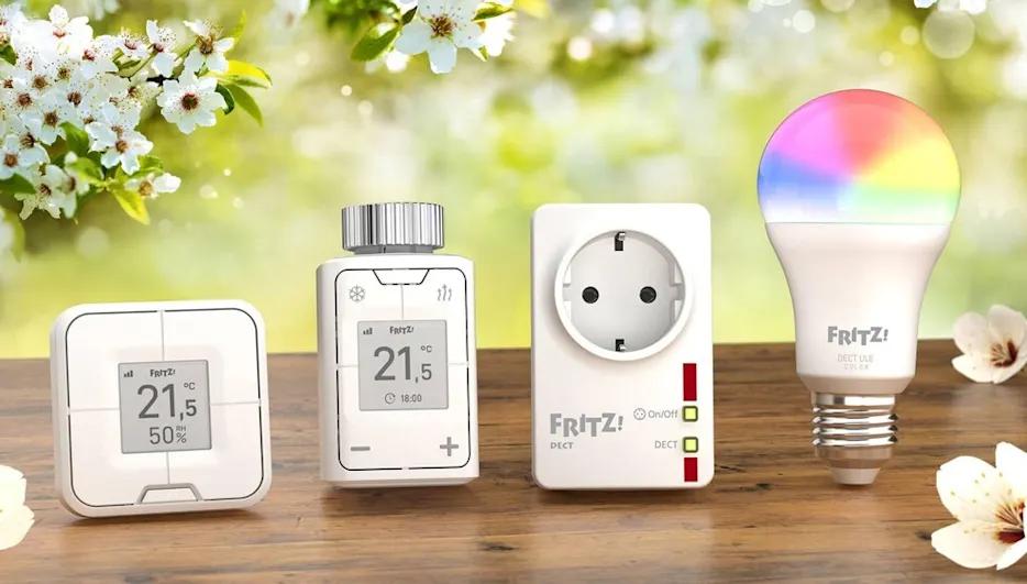Vier weiße FRITZ!-Smart-Home-Geräte, darunter Thermostate, eine Steckdose und eine Lampe, stehen auf einem Holztisch vor Blumen.