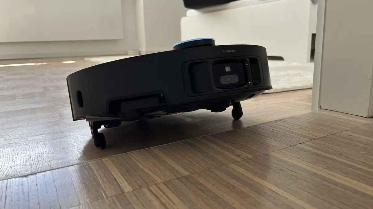 Ein schwarzer Staubsaugerroboter steht auf einem hellbraunen Holzboden. Im Hintergrund sind eine weiße Tür und ein weißes Möbelstück.