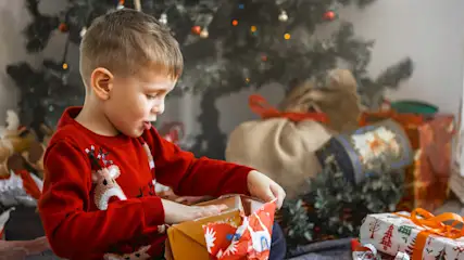 Ein Junge mit rotem Pullover öffnet ein Geschenk vor einem Weihnachtsbaum. Es gibt mehrere Geschenke im Hintergrund.