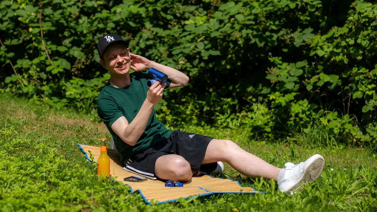 Alex sitzt im Park auf einer Strandmatte und kühlt sich mit dem Handventilator Shark ChillPill ab.