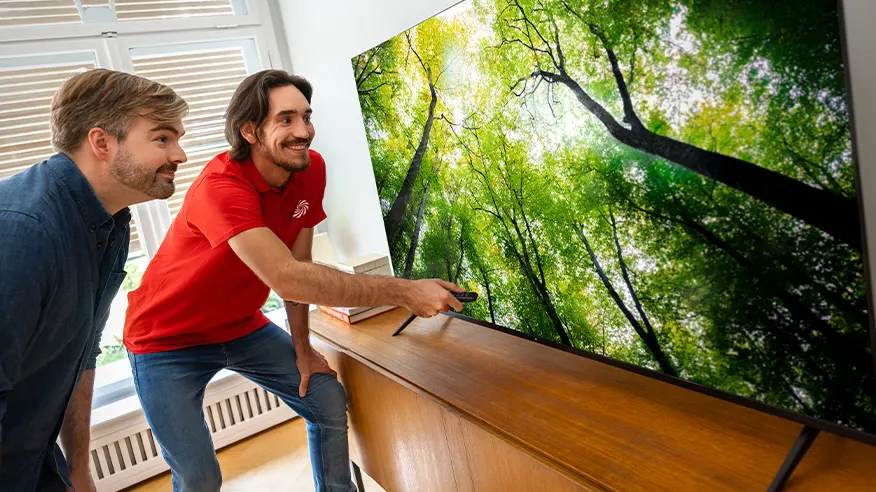 Dwóch mężczyzn ogląda telewizor pokazujący widok lasu. Jeden trzyma pilot. Telewizor stoi na drewnianej komodzie obok okna.