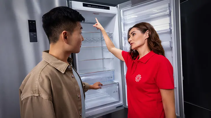 Kobieta sprzedawca w czerwonej koszulce stoi z mężczyzną przy otwartej lodówce i wskazuje na półki lodówki