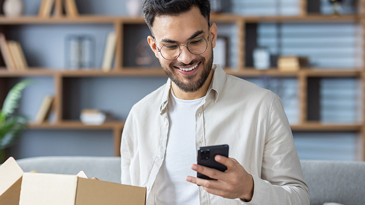 Mężczyzna w okularach uśmiecha się, patrząc na telefon. Obok otwarte kartonowe pudełko. W tle regał z książkami i roślinami.