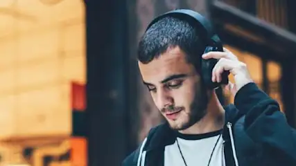 Mężczyzna w słuchawkach, ubrany w bluzę z kapturem, stoi na tle budynku z widocznym oświetleniem w tle.