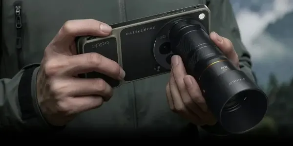 Zbliżenie: dwie dłonie trzymają smartfon OPPO z logo Hasselblad w poziomie; do aparatu zamocowany jest duży, cylindryczny obiektyw zewnętrzny; rozmyte tło plenerowe.