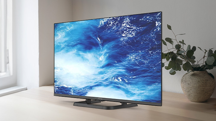 Telewizor z wyświetlanym obrazem wody stoi na stole obok rośliny w doniczce, w tle okno.