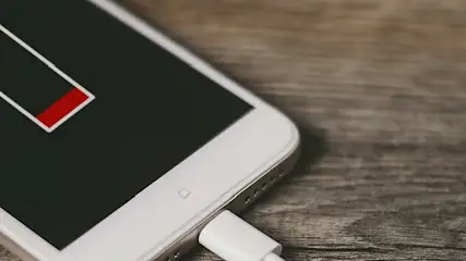 Na drewnianym stole leży biały smartfon z ikoną niskiego poziomu baterii i podłączonym kablem ładowarki.