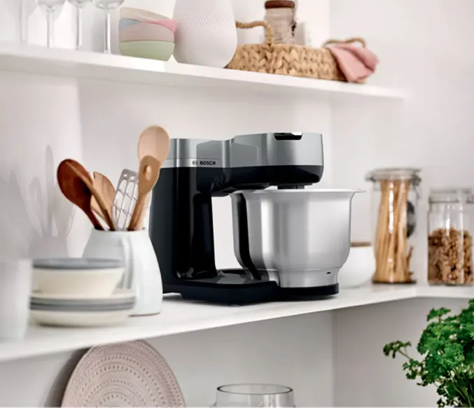 Na półce stoi czarno-srebrny robot kuchenny Bosch, obok miski z drewnianymi łyżkami. W tle słoiki z jedzeniem i zioła.