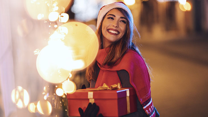 Kobieta trzymająca prezent na święta stojąc na dworze w czapce Mikołaja.