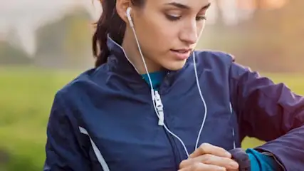 Kobieta w niebieskiej kurtce sportowej sprawdza zegarek; w tle zieleń i ścieżka.