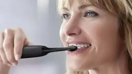 Kobieta myje zęby czarną, elektryczną szczoteczką. Tło jest jasne, rozmyte.