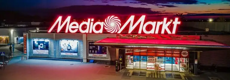 Oświetlony budynek Media Markt o zmierzchu. Nad wejściem neon z nazwą i logo sklepu. Szklane witryny z reklamami.