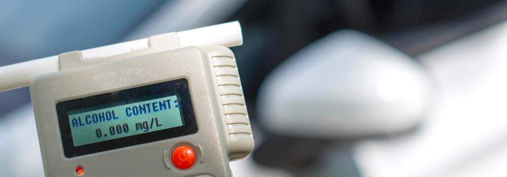Alkomat z wyświetlaczem wskazującym "ALCOHOL CONTENT: 0.000 mg/L", obok fragment samochodu.