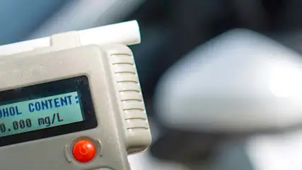 Alkomat z wyświetlaczem wskazującym "ALCOHOL CONTENT: 0.000 mg/L", obok fragment samochodu.