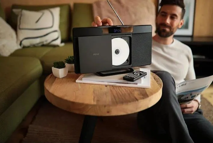 Mężczyzna siedzi obok okrągłego stolika z radiem, płytą CD, pilotem i magazynem w salonie z zieloną sofą.