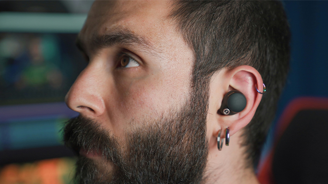 Mężczyzna z brodą, w uchu słuchawka bezprzewodowa i kolczyki w srebrnym kolorze. Tło jest rozmyte, widać fragment krzesła.