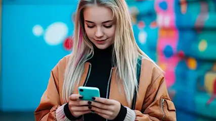 Kobieta używa telefonu na tle kolorowego graffiti. Nosi brązową kurtkę i czarny golf. Tło ma niebieskie, różowe i turkusowe akcenty.