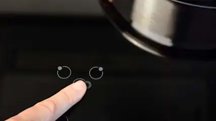 Palec wskazujący naciska przycisk na czarnej płycie indukcyjnej obok garnka. Widoczne symbole sterowania.