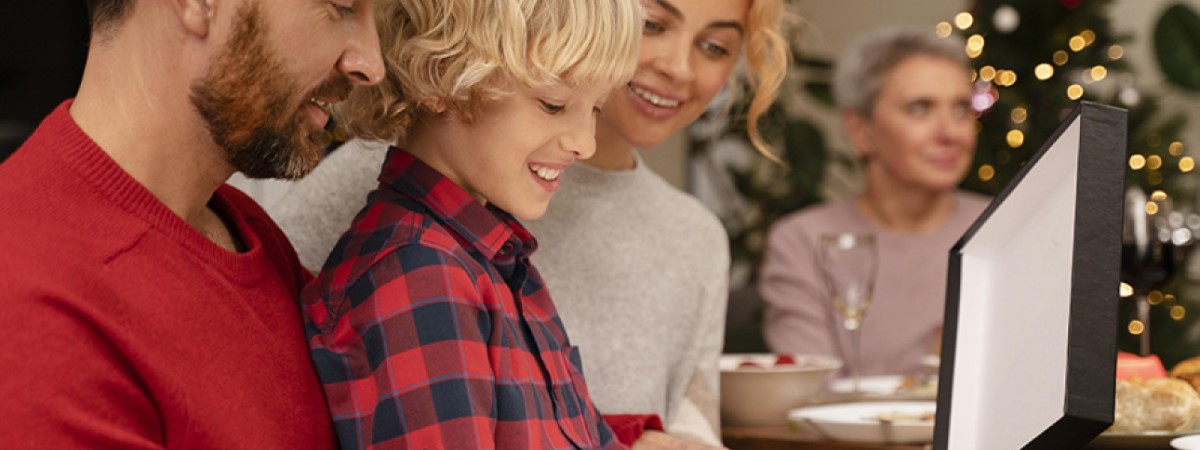 Rodzina patrzy na prezent. Mężczyzna w czerwonej koszuli, dziecko w kraciastej koszuli, kobieta w szarym swetrze i starsza kobieta. W tle choinka.