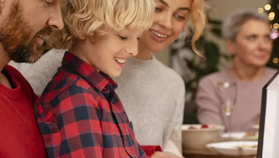Rodzina patrzy na prezent. Mężczyzna w czerwonej koszuli, dziecko w kraciastej koszuli, kobieta w szarym swetrze i starsza kobieta. W tle choinka.