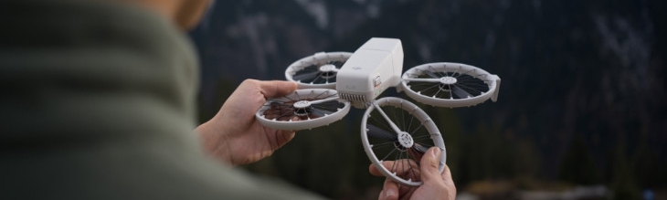Osoba trzyma białego drona z czterema śmigłami na tle gór i ciemnego tła.