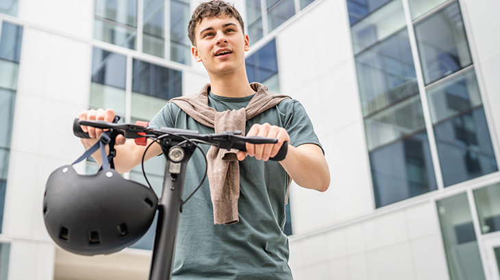 Zadowolony młody mężczyzna ze swetrem zawiązanym wokół szyi trzyma elektryczną hulajnogę, przez którą zawieszony jest kask ochronny.