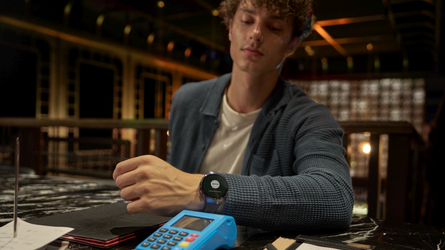 Mężczyzna patrzy na zegarek na ręku obok terminala płatniczego. W tle stolik, karta, długopis i elementy wystroju wnętrza.