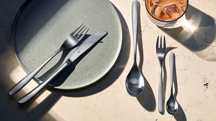 Na beżowym tle leżą talerz, sztućce i szklanka z napojem. Na talerzu leżą nóż i widelec. Obok talerza leżą łyżka, widelec i łyżeczka.