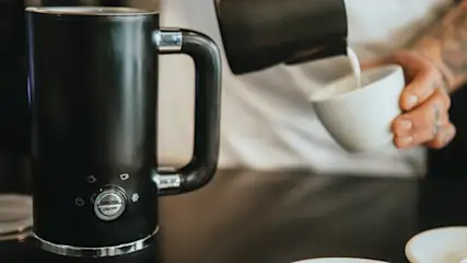 Osoba wlewa napój z czarnego dzbanka do białego kubka. Obok stoi czarny spieniacz do mleka.