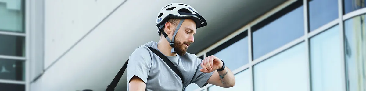 Mężczyzna w kasku rowerowym spogląda na zegarek na tle budynku z oknami.