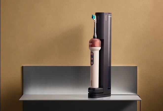 Szczoteczka elektryczna Dyson w beżowo-bordowym kolorze stoi w czarnej stacji ładującej na metalowym stoliku.