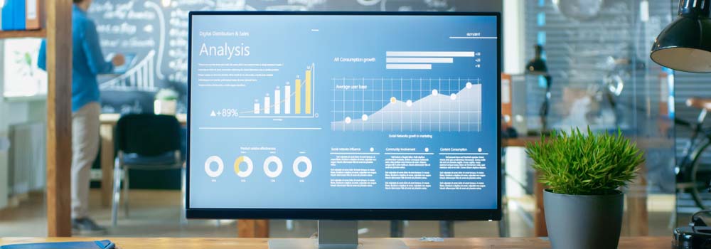 Monitor z wykresami i danymi na biurku, w tle roślina doniczkowa i osoba przy tablicy.