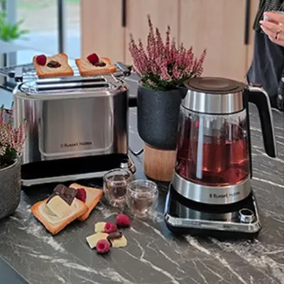 Na blacie stoi toster z grzankami, czajnik z napojem, szklanki, kwiaty w doniczce, tosty i maliny.