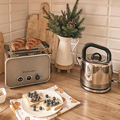 Na blacie kuchennym toster z pieczywem, czajnik, talerz z tostami i borówkami. W tle deska do krojenia i wazon z gałązkami.