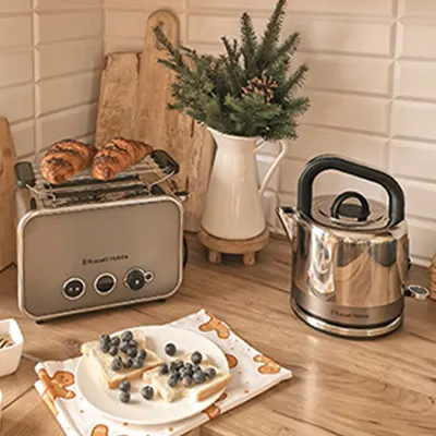 Na blacie kuchennym toster z pieczywem, czajnik, talerz z tostami i borówkami. W tle deska do krojenia i wazon z gałązkami.