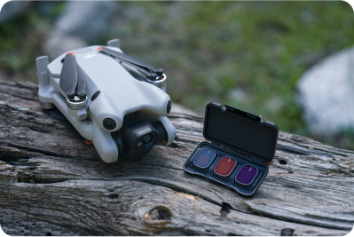 Na teksturowanym drewnie leży szary dron obok otwartego czarnego pudełka z filtrami: niebieskim, czerwonym i fioletowym. Tło rozmyte, zielone.