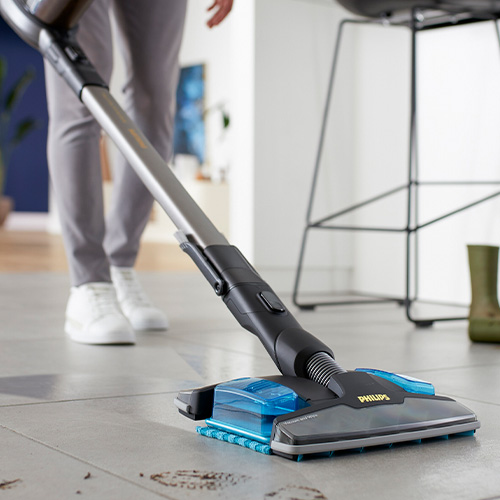 Osoba odkurza szarą podłogę odkurzaczem Philips. W tle widoczne krzesło barowe i zielone buty.