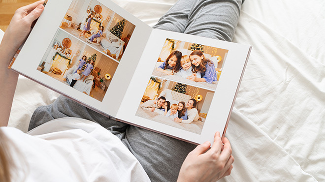 Osoba w szarych spodniach i białej koszuli trzyma otwarty album ze zdjęciami rodzinnymi na białym łóżku.