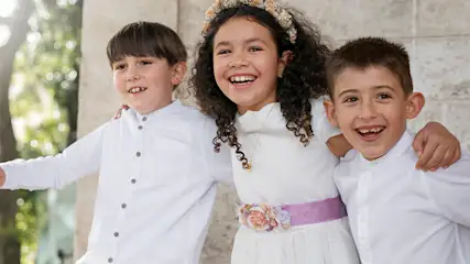 Troje dzieci w białych ubraniach, uśmiechniętych, stoi blisko siebie na tle zieleni i jasnych budynków.