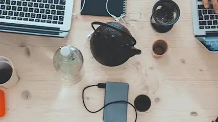 Na drewnianym stole leżą laptopy, notatniki, czajnik, kubki, miska z przekąskami, telefon i przenośny dysk.