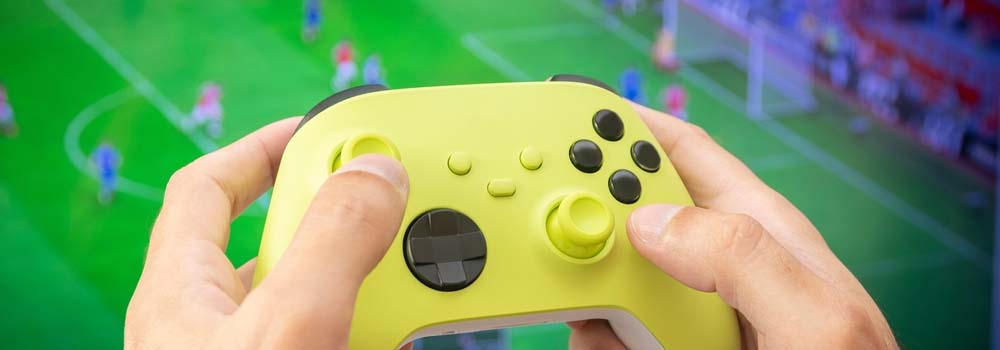 Dłonie trzymają żółty kontroler do gier. W tle widoczny zielony ekran z postaciami w ruchu, przypominający grę sportową.