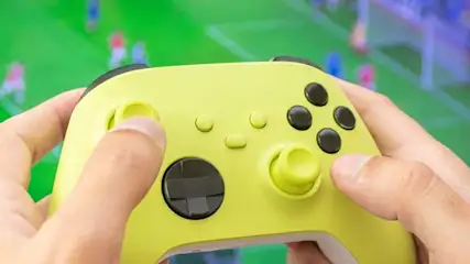 Dłonie trzymają żółty kontroler do gier. W tle widoczny zielony ekran z postaciami w ruchu, przypominający grę sportową.