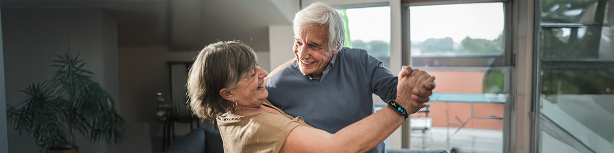 Para tańcząca w jasnym pokoju, widoczna roślina doniczkowa i okno z widokiem na zewnątrz.