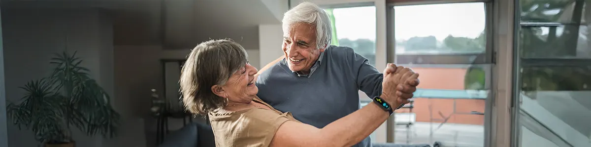Para tańcząca w jasnym pokoju, widoczna roślina doniczkowa i okno z widokiem na zewnątrz.