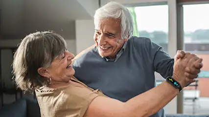 Para tańcząca w jasnym pokoju, widoczna roślina doniczkowa i okno z widokiem na zewnątrz.
