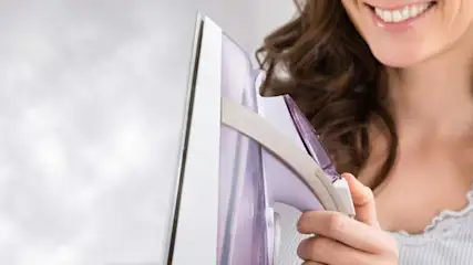 Kobieta prasuje fioletową koszulę na desce do prasowania w jasnym pokoju. Uśmiecha się, trzymając żelazko w dłoni.