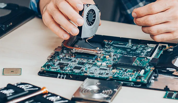 Osoba naprawia płytę główną laptopa, trzyma wentylator. Obok dysk twardy, procesor i narzędzia.