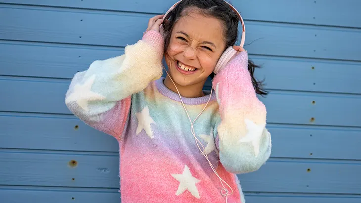 Uśmiechnięta dziewczyna w słuchawkach i kolorowym swetrze ze wzorem gwiazd stoi na tle niebieskiej drewnianej ściany.