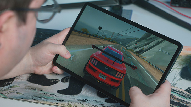 Osoba trzyma tablet z grą wyścigową. Na ekranie czerwony samochód na drodze, w tle góry. Tablet leży na podkładce z wzorem.