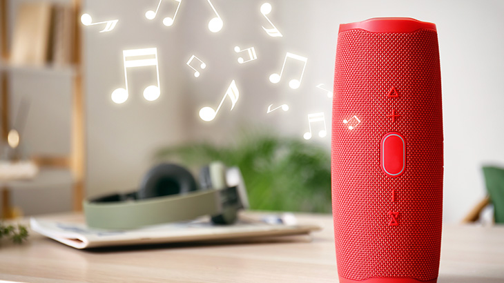 Na stole stoi czerwony głośnik. W tle widać słuchawki i nuty muzyczne.